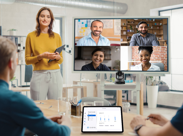 Image of a conference with a woman standing next to a display featuring 4 individuals, with a Q-SYS network camera below.